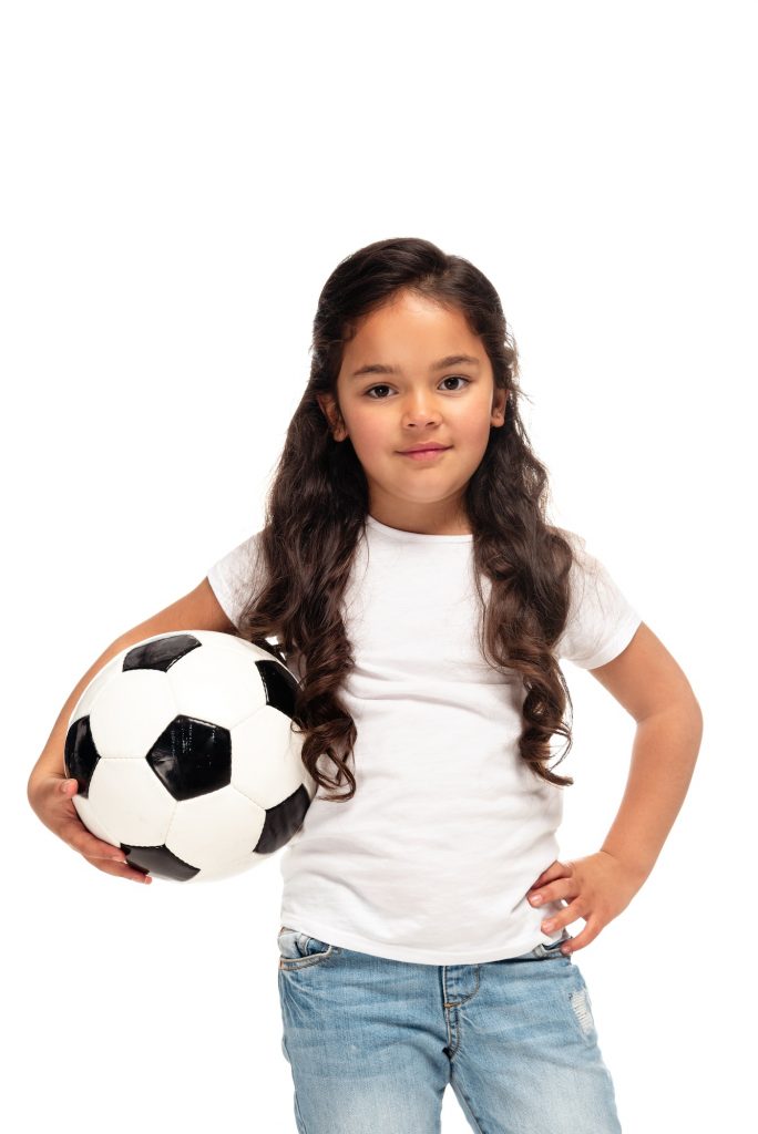 cheerful latin kid standing with hand in hip and holding soccer ball isolated on white